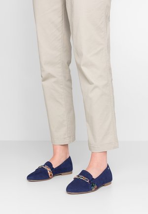 Marineblå ruskinds loafers med en multicolor flettet kædedetalje og flad sål, parret med lysebeige cropped bukser.