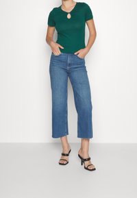 Vihreä lyhythihainen yläosa, jossa on avainreikäyksityiskohta, yhdistetty korkeavyötäröisiin sinisiin leveälahkeisiin farkkuihin ja mustiin korollisiin sandaaleihin neutraalilla taustalla.