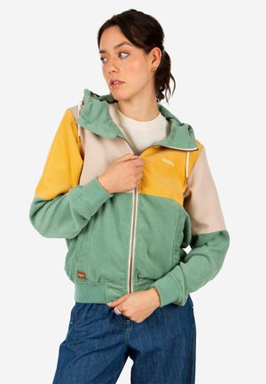 Woman wearing zip-up hoodie with color blocks in green, yellow, and beige, paired with blue jeans, looking to her left.