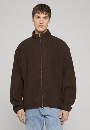 Młody mężczyzna ubrany w ciemnobrązową polarową bluzę na zamek i jasnoniebieskie dżinsy, stojący na tle jednolitego szarego tła.