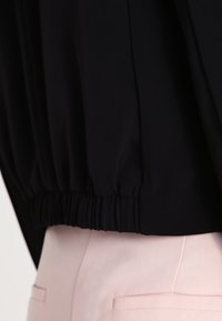 Vêtement noir avec une taille élastique, présentant une texture douce. Porté par-dessus un tissu rose clair, affichant des couleurs contrastées et une coupe décontractée.