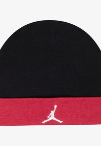 Mustast kootud müts punase alumise äärega, millel on valge Jumpmani logo. Sile tekstuur, ümardatud tipp ja kontrastne värviskeem.