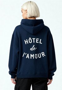 Felpa blu navy con grande testo bianco "HÔTEL de l'AMOUR" sul retro, realizzata in tessuto morbido, dotata di cappuccio con cordino e polsini a costine.