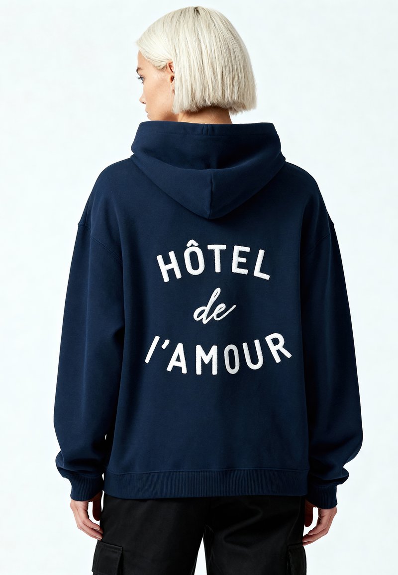 Felpa blu navy con grande testo bianco "HÔTEL de l'AMOUR" sul retro, realizzata in tessuto morbido, dotata di cappuccio con cordino e polsini a costine.