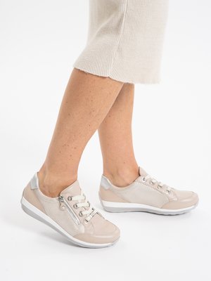 Beine mit beigen Sneakers in niedriger Ausführung, weißen Schnürsenkeln, seitlichem Reißverschluss und weißen Sohlen, kombiniert mit einem beigen Strickrock vor weißem Hintergrund.