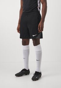 Schwarze Nike-Fußballshorts mit einem elastischen Bund, kombiniert mit weißen, knielangen Socken mit einem schwarzen Logo und schwarzen Fußballschuhen.