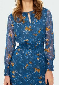 Robe florale bleue avec de longues manches transparentes, présentant un décolleté en forme de trou de serrure et une taille froncée, rehaussée de fleurs orange et blanches.