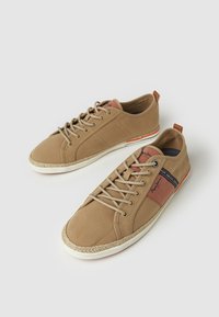 Zapatillas de lona beige con puntera redondeada, acentos de cuero marrón, diseño de cordones y suela trenzada con un detalle de franja multicolor.