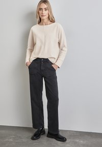 Beige strukturierter Pullover mit weitem Ausschnitt, kombiniert mit hochgeschnittenen, schwarzen weit geschnittenen Jeans und schwarzen Schnürschuhen.