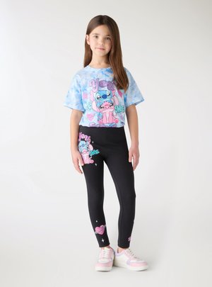 Młoda dziewczyna stoi, ubrana w niebieski t-shirt z nadrukiem kreskówki i czarne legginsy z pasującymi wzorami kreskówkowymi oraz różowo-białe trampki.