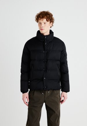 Jachetă puffer neagră cu guler înalt, având secțiuni orizontale quilted, nasturi prin apăsare și buzunare cu fermoar pe laterale. Purtați cu pantaloni verde închis.
