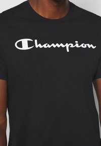 Černé bavlněné tričko s bílým logem "Champion" v kurzívě. Standardní provedení s kulatým výstřihem a krátkými rukávy. Hladká textura.