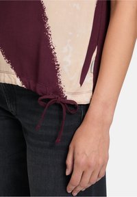 Nahaufnahme einer Hand, die neben dem Körper ruht, getragen von einem beige- und burgunderfarbenen Oberteil mit gebundenem Kordelzug-Saum und dunklen Jeans.