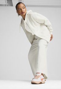 Giovane donna inclinata in avanti, con felpa bianca, pantaloni larghi e sneakers Puma bianche e rosa, su sfondo bianco uniforme.