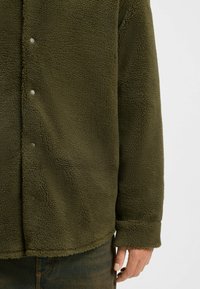 Olivgrüne Fleecejacke mit weicher, strukturierten Oberfläche, ausgestattet mit Druckknöpfen und einem lässigen Schnitt, der bis zur Hüfte reicht.