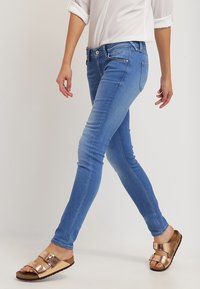 Úzké modré džíny z denimu s pasem ve střední výšce, pěti kapsami a vybledlým vzhledem. Kombinovány s hnědými sandály na plochém podpatku.