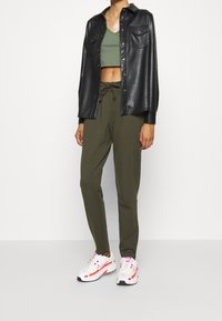 Persona che indossa una giacca di pelle nera sopra un top corto verde e pantaloni con cordino verde oliva, abbinati a sneakers Nike bianche e rosa.