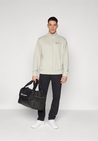 Grå zip-up jacka med liten logotyp, kombinerad med mörka byxor. Personen håller en svart duffelbaga med logotyp och nätaccent.