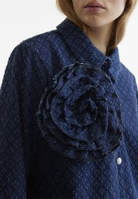 Camicia in tessuto testurizzato blu navy con un ampio appliqué floreale a strati sul lato sinistro e un piccolo dettaglio con bottone bianco sul davanti.