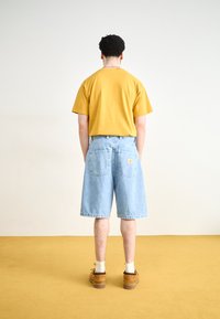 Tricou din bumbac galben, croi lejer; pantaloni scurți din denim albastru deschis cu buzunare în spate; pantofi boat maro cu șosete albe; fundal simplu.