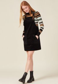 Musta corduroy-henkari mekko, jossa on kaksi etupoketta, säädettävät olkahihnat ja suora leikkaus. Päälle puettuna raidallinen pitkähiha-tyyli.