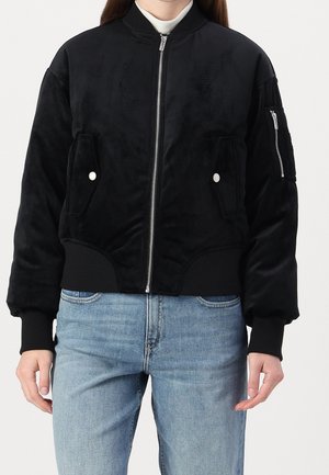 Blouson bomber noir à fermeture éclair avec poches avant à rabat et poche zippée sur la manche, porté sur un col roulé blanc et un jean bleu clair.