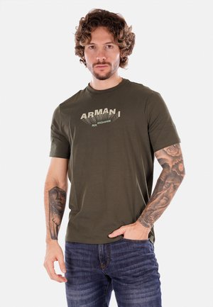 T-shirt in cotone verde oliva con maniche corte, caratterizzata da una stampa grafica con la scritta "ARMAN AIX EXCHANGE" in beige chiaro sul petto.
