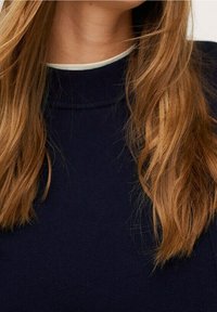 Lunghi capelli castano chiaro e ondulati che cadono su un maglione a girocollo blu navy con un colletto bianco sottostante.