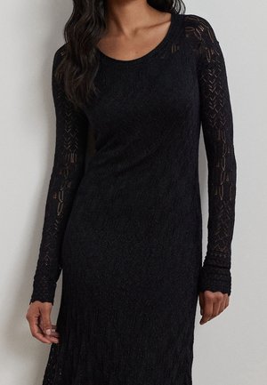 Kvinde iført en langærmet sort blondekjole med rund halsudskæring, stående foran en ensfarvet lys baggrund.