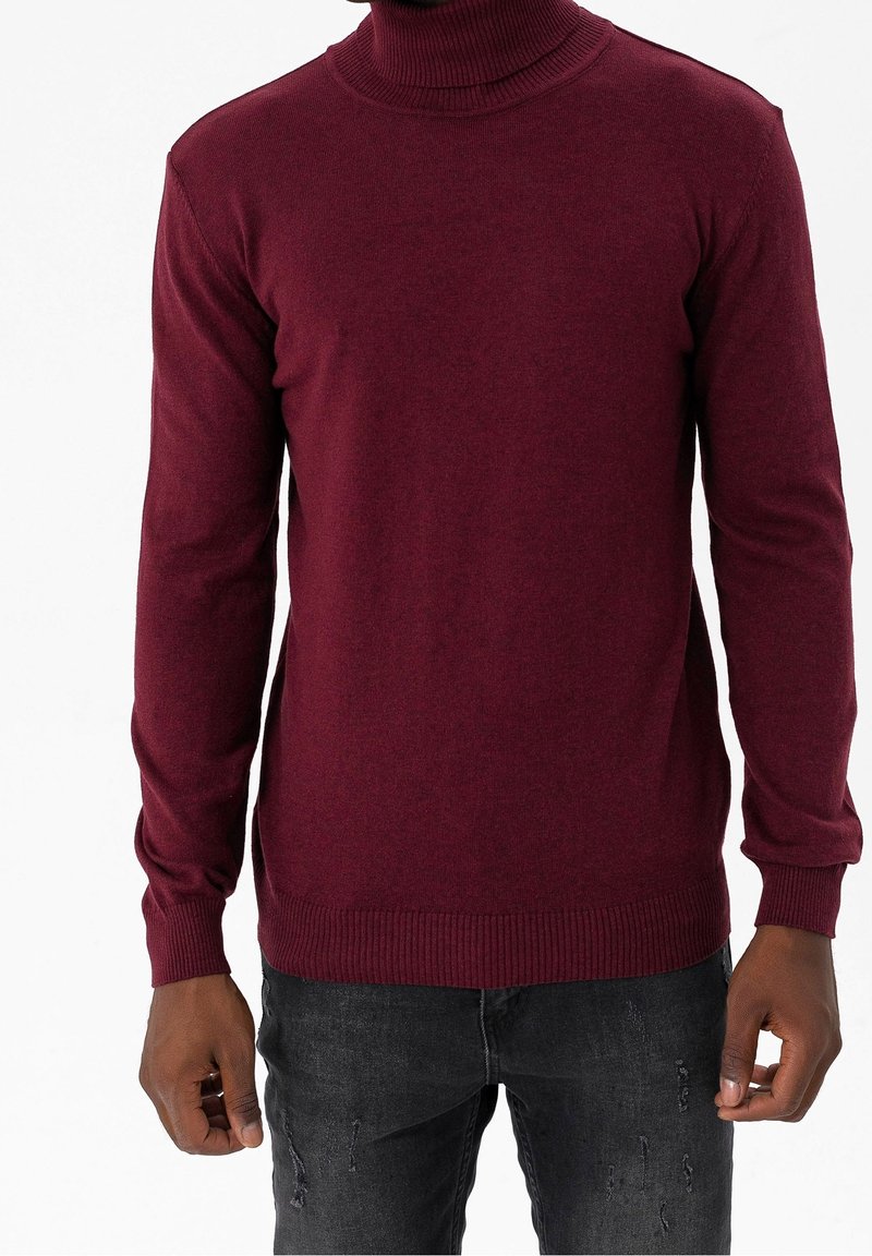 Jersey de cuello alto burdeos con mangas largas, textura acanalada y dobladillo ajustado, combinado con jeans oscuros desgastados.