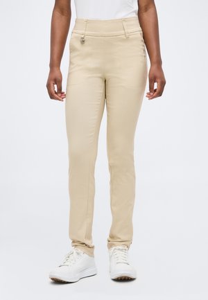 Persona che indossa pantaloni beige slim con dettaglio a bottone e sneakers bianche, in piedi su uno sfondo chiaro e uniforme.
