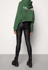 Persona che indossa leggings in pelle sintetica neri, stivali platform neri e un hoodie verde corto con testo bianco sulla schiena, in piedi contro un muro semplice.