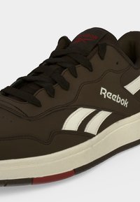 Καφέ αθλητικό παπούτσι Reebok με μαύρα κορδόνια, λευκές πλαϊνές ρίγες και κόκκινο λογότυπο στη γλώσσα, σε κοντινό πλάνο μπροστά σε απλό φόντο.