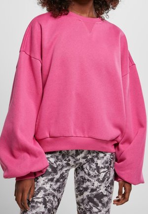 Osoba ubrana w oversize'ową, jasnoróżową bluzę z opuszczonymi ramionami i balonowymi rękawami, zestawioną z czarno-białymi legginsami w wzory.