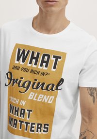 Valge puuvillane T-särk, millel on julged musta ja kuldse kollase värviga trükitud tekstid. Sõnumid sisaldavad "WHAT," "Original Blend" ja "WHAT MATTERS."