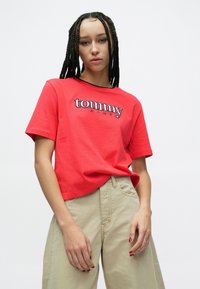 Raudonas medvilninis marškinėlių trumpomis rankovėmis, puoštas siuvinėjimu „tommy“ ir „JEANS“ kontrastingomis spalvomis. Derinamas su smėlio spalvos plačiomis kelnėmis.