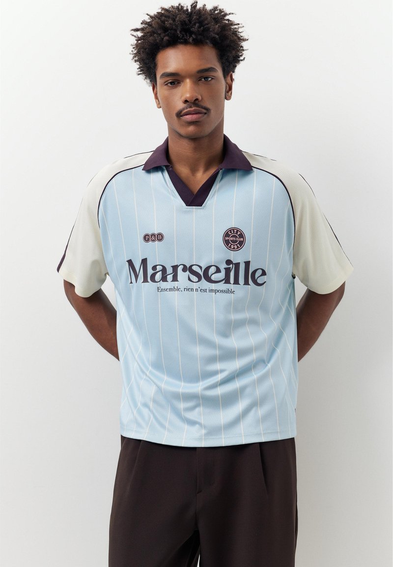 Junger Mann trägt ein hellblau-weißes Sporttrikot mit den Logos "Marseille" und "City Sports Club 1991" und steht vor einem schlichten Hintergrund.