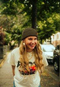 Warm olive beanie, cream graphic t-shirt with a pixelated car design, layered gold necklaces, long wavy hair with braids, outdoor setting.