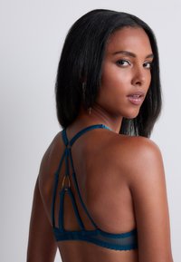 Bralette en dentelle teal avec un dos strappy, présentant des anneaux décoratifs et des bretelles ajustables. Tissu doux et texturé avec des détails sophistiqués.