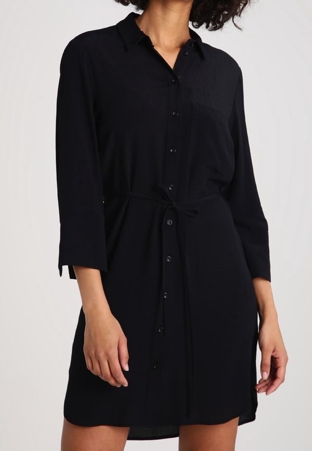 Svart skjortklänning i lätt tyg, med knapplist fram, krage och ett knytbälte i midjan. Långa ärmar med uppvikt ärmslut.