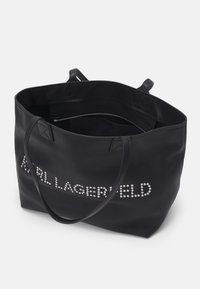 Sac cabas en cuir noir avec des anses fines et texte clouté argenté sur le devant, ouverture supérieure montrant une petite poche intérieure zippée.
