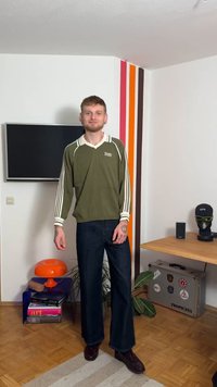 Alyvuogių žalios spalvos megztinis su v formos iškirpte, baltais akcentais ir trimis baltomis juostomis ant rankovių, derinamas su tamsių džinsų išplatėjusiais kelnėmis ir rudo spalvos batais.