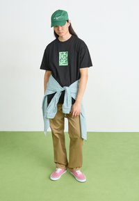 T-shirt noire avec un graphique vert, chemise rayée bleu clair nouée à la taille, pantalon beige et baskets roses sur un sol vert.