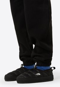 Mustad tikandiga sussid kergelt kummi tallaga, millel on küljele paigaldatud logo ning mida kannavad mustad spordipüksid ja sinised sokid.