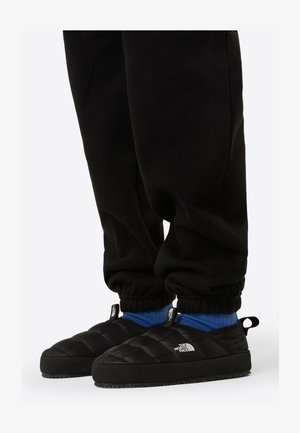 Chaussons noirs matelassés à enfiler avec une semelle en caoutchouc, ornés d'un logo sur le côté, portés avec un pantalon de jogging noir et des chaussettes bleues.