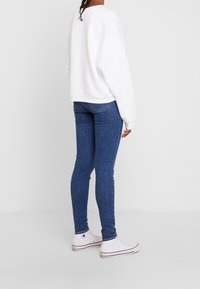 Bílá oversized mikina, modré skinny džíny a bílé tenisky. Přiléhavý design, hladká textura, s viditelnými detaily šití na džínech.