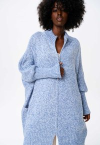 Lichtblauw gebreid trui met een hoge kraag, lange, brede mouwen en een losse pasvorm. Heeft een halve ritsluiting en ribgebreide accenten.