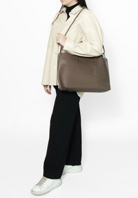 Bruine leren totebag met dubbele handvatten, rechthoekige vorm, minimaal ontwerp en een afneembare tag. Gepaarde met een crème shirt en zwarte broeken.