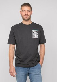 T-shirt en coton noir avec un imprimé graphique sur la partie gauche de la poche poitrine, et un col rond. Porté avec un jean en denim bleu clair.