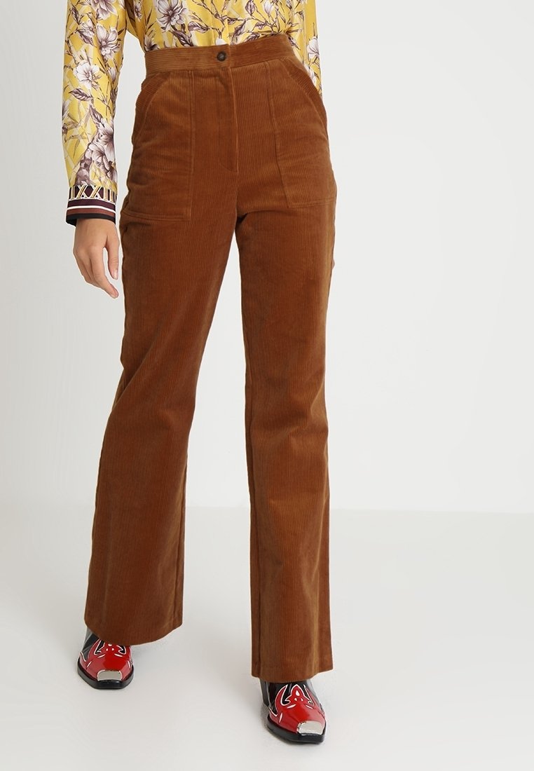 Personne portant un pantalon en velours côtelé marron taille haute, une chemise jaune à manches longues à motifs floraux, et des chaussures rouges brillantes à bout pointu, debout sur un sol blanc.